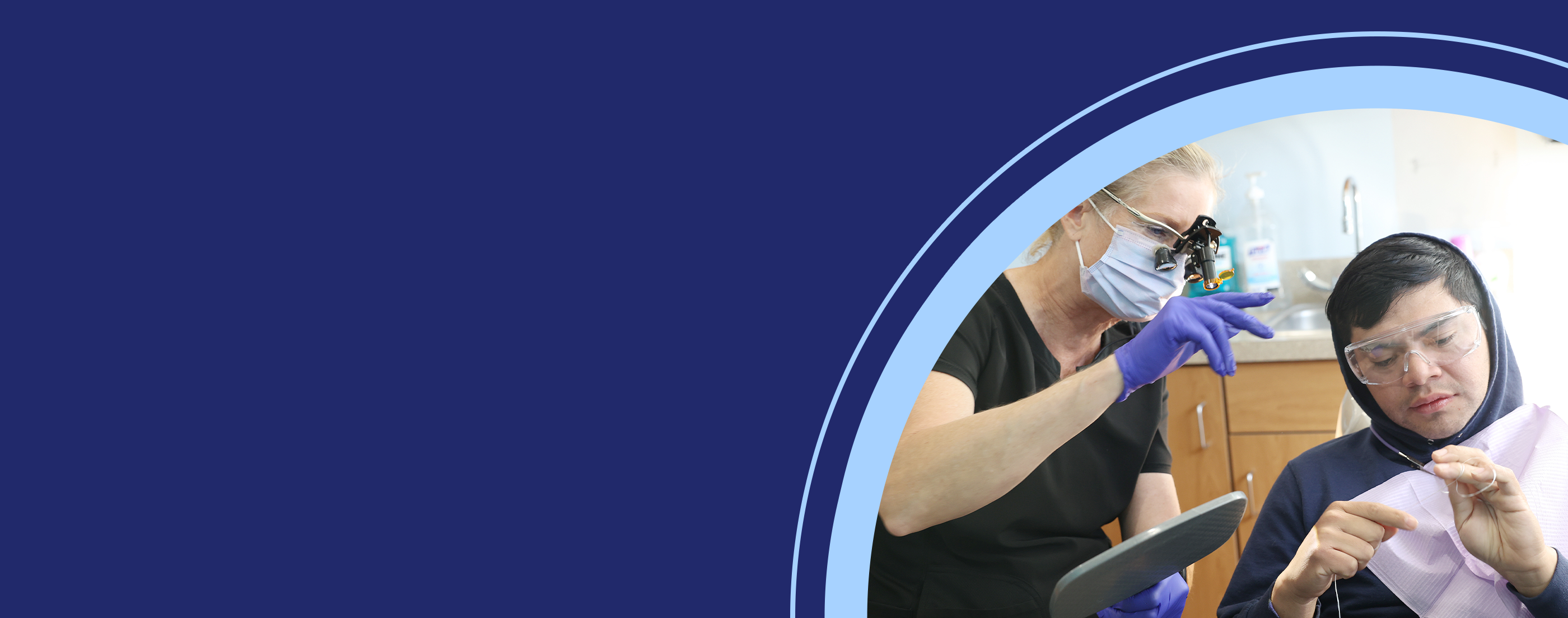 A dental provider wearing magnifying loupes and gloves examines dental floss with a patient seated in a clinic chair