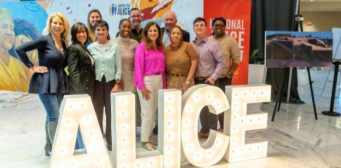 A group of National ALICE Summit 2026 attendees stand and smile behind giant light-up ALICE letters, with a vibrant mural in the background