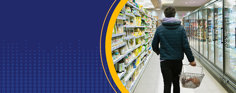 A person in a dark blue, puffy hooded jacket walks away from the camera and down a supermarket aisle with household items stacked on the shelves on both sides. The person carries a shopping basket containing only the few items they could afford as they walk toward the checkout lanes.