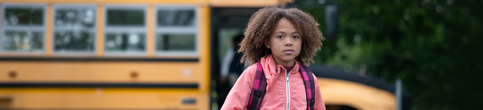 A school-age child, wearing a backpack and a pink jacket, stands in front of a school bus and stares at the camera with a serious expression.
