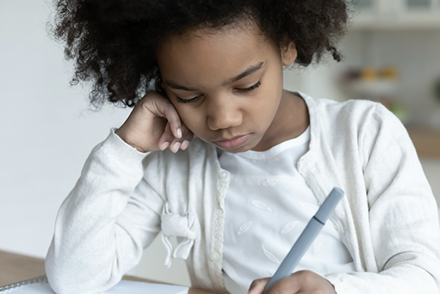 Young student sitting at a desk, looking down, and writing with a pen on paper